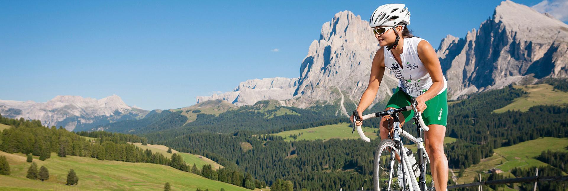 A woman on a racing bicycle riding along a mountain road with a view of the Dolomites.